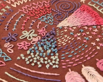May include: Close-up of a brown fabric with colorful embroidery. The embroidery features various stitches in shades of pink, blue, purple, and white, including flowers, spirals, and geometric patterns. The craftwork showcases a variety of textures and designs.