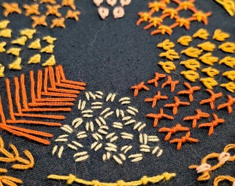 May include: Close-up of a dark blue fabric with various embroidery stitches in shades of orange, yellow, and cream. The embroidery includes geometric patterns, lines, and textured designs, showcasing a handcrafted textile art piece.
