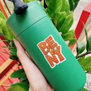 May include: A green insulated tumbler with a black lid. The tumbler has a sticker that says "BE NY" in orange letters with white dots. The sticker also has a small green design.