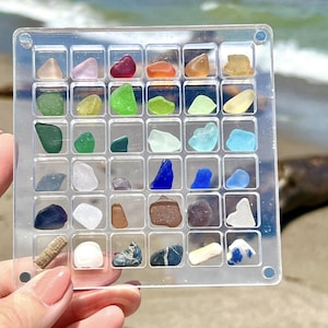 Puede incluir: Una rejilla de plástico transparente llena de varios trozos de vidrio de mar de colores. Los fragmentos de vidrio de mar son en tonos de verde, azul, rojo, naranja y blanco. La rejilla se sostiene contra un fondo de playa con agua y árboles.