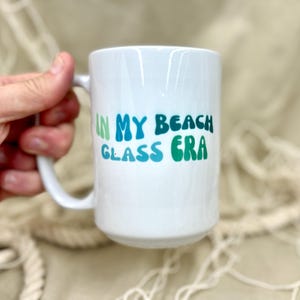 May include: White ceramic mug with a green, blue and light green design that says "In My Beach Glass Era".