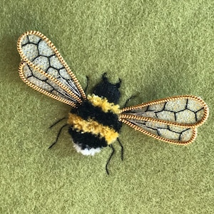 May include: A handmade bee brooch with a fluffy yellow and black body, delicate wire-framed wings, and small antennae. The wings feature a honeycomb design. The brooch is set against a green background.