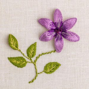 May include: Embroidered purple flower with six petals and a green stem and leaves. The flower has white and purple detailing. The leaves are green with yellow accents. The embroidery is on a white fabric background.