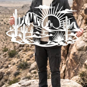 A person standing outdoors on rocks holds up a white metal laser-cut desert sunset sign with cacti.