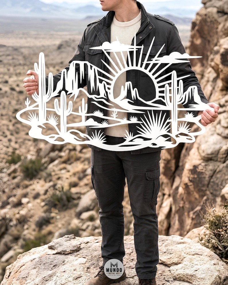 A person standing outdoors on rocks holds up a white metal laser-cut desert sunset sign with cacti.