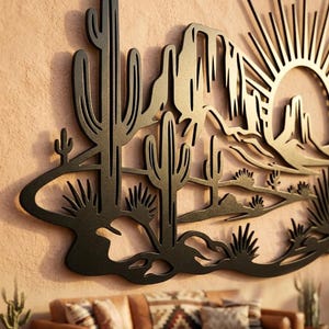 A black metal laser-cut wall art piece depicting a desert landscape with saguaro cacti, mountains, and a rising sun mounted on a textured beige wall.