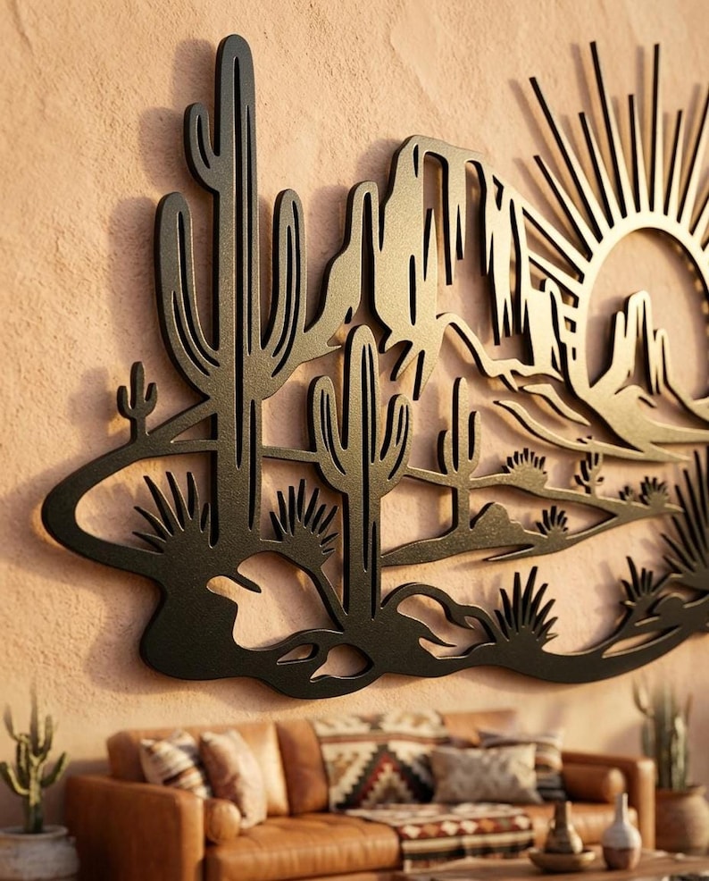 A black metal laser-cut wall art piece depicting a desert landscape with saguaro cacti, mountains, and a rising sun mounted on a textured beige wall.