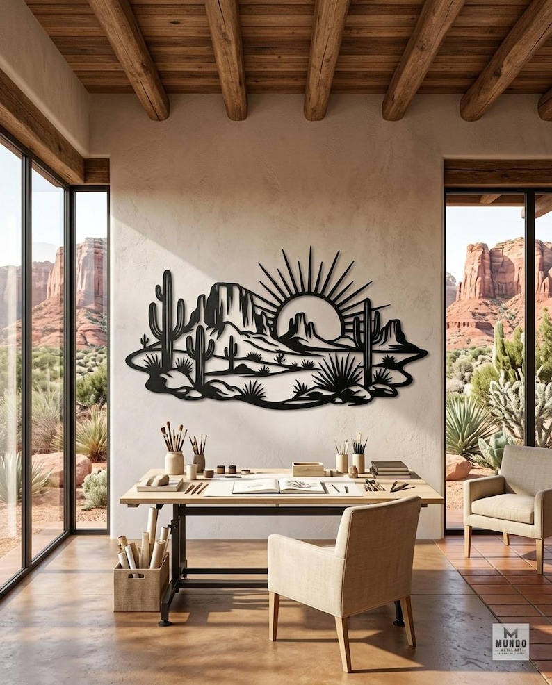 A black metal desert sunset wall hanging is mounted in a bright studio workspace overlooking red rock formations.