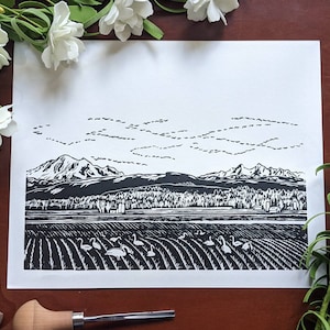 Könnte beinhalten: Schwarz-weißer Linolschnitt mit einer Bergkette und einem Feld mit Gänsen im Vordergrund. Der Druck ist auf weißem Papier und hat ein rustikales, handgemachtes Aussehen.