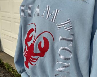 May include: Light blue sweatshirt with the word "HAMPTONS" embroidered in white and blue striped letters. A red lobster graphic is on the front. The sweatshirt is a casual style.