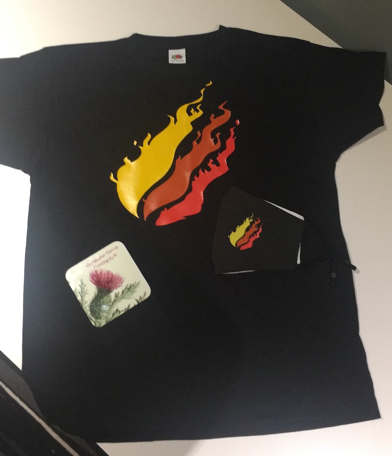 May include: A black t-shirt with a yellow, orange, and red flame design. The shirt is laying flat on a white surface. There is a small black square with a smaller flame design on it and a square coaster with a purple flower on it.
