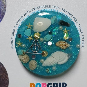 May include: A blue and white PopGrip phone stand with a Harry Potter symbol and seashells. The text "POPSOCKETS" and "POPGRIP" are on the packaging. The text "PHONE GRIP & STAND WITH SWAPPABLE TOP. TRY ME! PULL TWICE TO GRIP." is on the packaging.