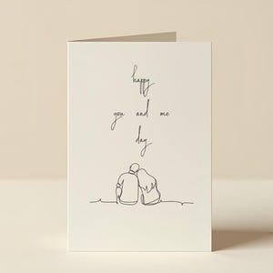 May include: A folded greeting card with a minimalist line drawing of two figures embracing. The card features the handwritten text "happy you and me day" in black ink on a cream background.