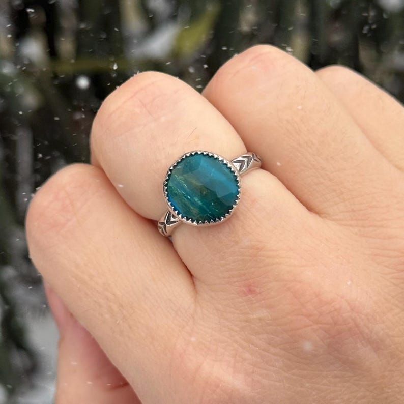 Pu&ograve; includere: Un anello d'argento con una grande gemma ovale color teal incastonata in una lunetta smerlata. La fascia dell'anello presenta un motivo a freccia su entrambi i lati della gemma. L'anello &egrave; mostrato su un dito.