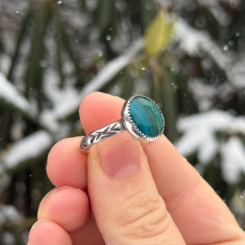 Pu&ograve; includere: Un anello d'argento con una grande pietra rotonda color teal. L'anello ha una fascia strutturata con un motivo a spina di pesce. La pietra &egrave; incastonata in una lunetta con un bordo smerlato. Lo sfondo &egrave; sfocato con la neve.