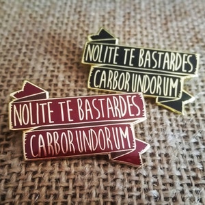 Puede incluir: Dos pines de esmalte de color dorado con un diseño de bandera roja y negra. El texto en los pines dice "Nolite Te Bastardes Carbor Undorum".