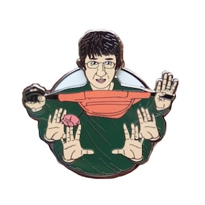 Puede incluir: Un pin de esmalte verde con una ilustración de dibujos animados de una persona que lleva una riñonera roja y sostiene un pequeño objeto rosa en la mano.