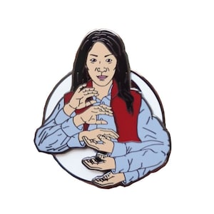 Puede incluir: Pin esmaltado con una ilustración estilizada de una persona con múltiples brazos. La persona lleva un chaleco rojo sobre una camisa azul claro. El pin está sobre un fondo blanco con un borde negro.