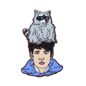 Puede incluir: Un pin de esmalte azul con una ilustración de dibujos animados de una persona que lleva una chaqueta azul con un mapache en la cabeza.