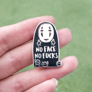 May include: A black and silver enamel pin featuring a character from Spirited Away with the text "NO FACE NO FUCKS". The pin has a white face and small cartoon figures at the bottom.