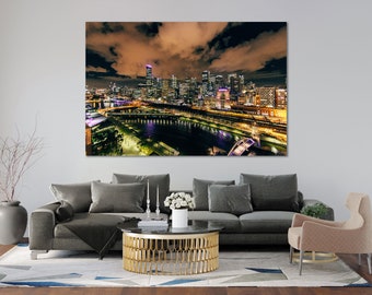 May include: A canvas print of a cityscape at night, showcasing illuminated skyscrapers and a river. The image features warm lighting from buildings and streetlights, contrasted by dark clouds. The artwork is displayed above a gray sofa.