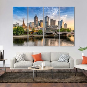 May include: A panoramic view of the Melbourne skyline with a bridge over a river in the foreground. The city skyline features tall buildings, including a church with a spire. The sky is a light blue with some clouds.