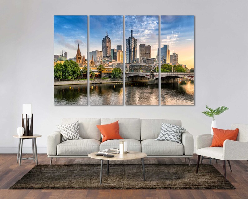 May include: A panoramic view of the Melbourne skyline with a bridge over a river in the foreground. The city skyline features tall buildings, including a church with a spire. The sky is a light blue with some clouds.