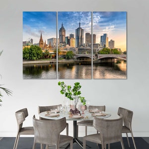 May include: A panoramic view of the Melbourne skyline with a bridge over a river. The city is filled with tall buildings, including a church with a tall spire. The sky is blue with white clouds.