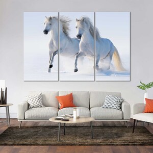 May include: Two white horses run through a snowy field, their manes flowing in the wind.