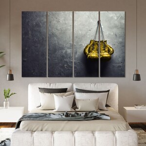 May include: Two gold boxing gloves hanging on a gray textured wall. The gloves are tied together with a black cord.