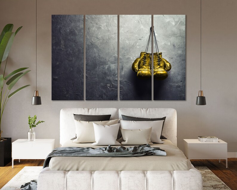 May include: Two gold boxing gloves hanging on a gray textured wall. The gloves are tied together with a black cord.