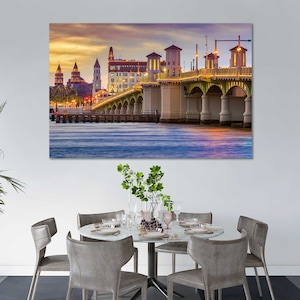 May include: A photograph of a bridge over a body of water with buildings in the background. The bridge is lit up at night and the water is reflecting the lights. The buildings are also lit up and there are trees in the foreground.