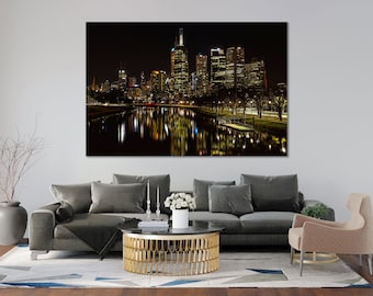 May include: A large canvas print of a nighttime cityscape. The image features a river reflecting the illuminated buildings and lights. The buildings are tall and modern, with many windows. The overall color palette is dark, with warm yellow and orange lights.