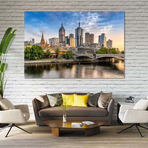 May include: A panoramic view of the Melbourne skyline with a bridge in the foreground and a river reflecting the city buildings. The sky is blue with white clouds and the sun is setting.