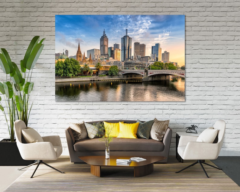 May include: A panoramic view of the Melbourne skyline with a bridge in the foreground and a river reflecting the city buildings. The sky is blue with white clouds and the sun is setting.