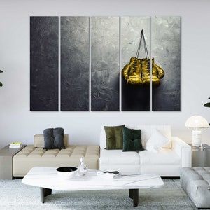 May include: A pair of gold boxing gloves hanging on a gray textured wall. The gloves are tied together with a black cord.