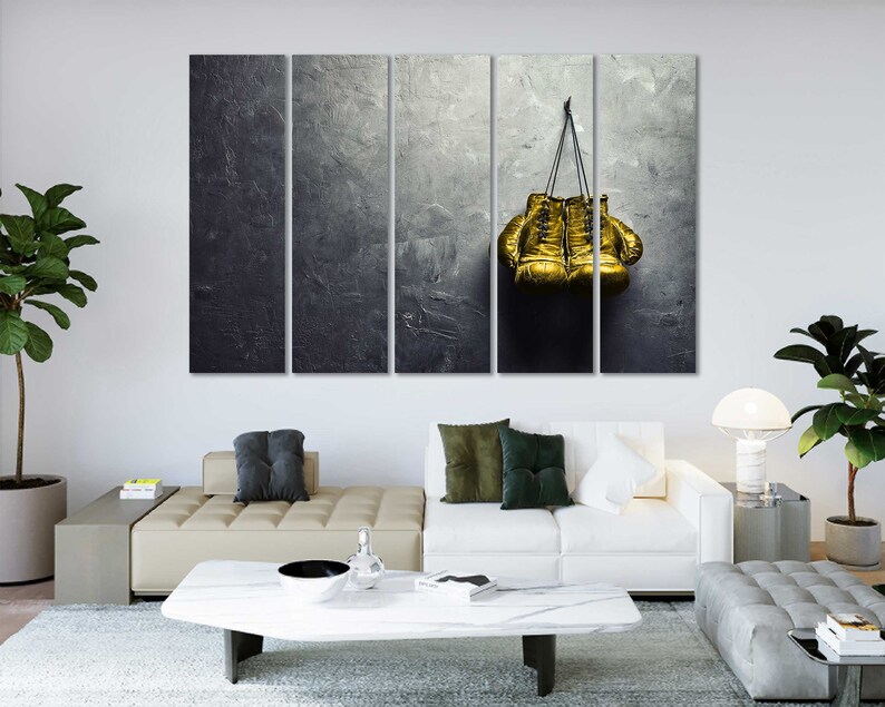 May include: A pair of gold boxing gloves hanging on a gray textured wall. The gloves are tied together with a black cord.