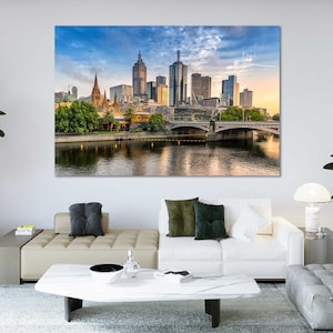 May include: A panoramic view of the Melbourne skyline at sunset, featuring a bridge over a river and tall buildings. The sky is a mix of blue and orange, with clouds.