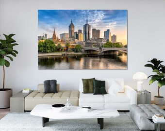 May include: A panoramic view of the Melbourne skyline at sunset, featuring a bridge over a river and tall buildings. The sky is a mix of blue and orange, with clouds.