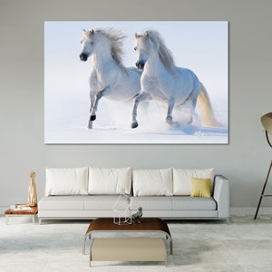 May include: Two white horses running through a snowy field. The horses are running towards the right side of the image. The snow is flying up behind them.