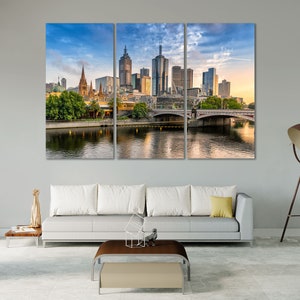 May include: A panoramic view of the Melbourne skyline with a bridge in the foreground. The city is filled with tall buildings, including a church with a spire. The sky is blue with white clouds. The water is calm and reflects the buildings.