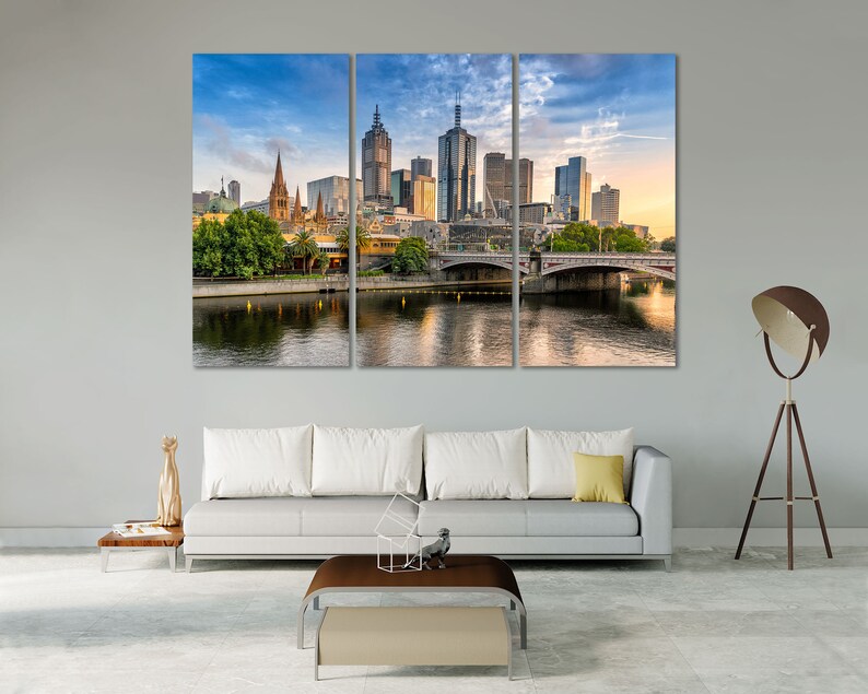 May include: A panoramic view of the Melbourne skyline with a bridge in the foreground. The city is filled with tall buildings, including a church with a spire. The sky is blue with white clouds. The water is calm and reflects the buildings.