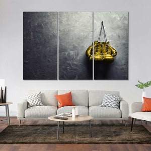 May include: A pair of gold boxing gloves hanging on a gray textured wall. The gloves are tied together with a black cord.
