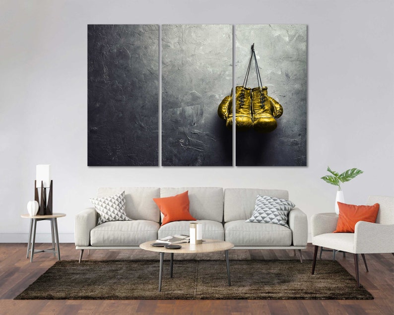 May include: A pair of gold boxing gloves hanging on a gray textured wall. The gloves are tied together with a black cord.