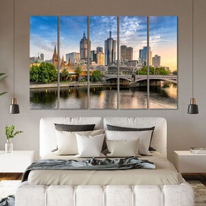 May include: A panoramic view of the Melbourne skyline with a bridge in the foreground. The city is bathed in the warm glow of the setting sun, with tall buildings and a church steeple visible. The river reflects the sky and buildings, creating a serene and picturesque scene.