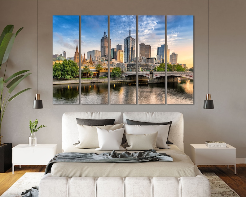 May include: A panoramic view of the Melbourne skyline with a bridge in the foreground. The city is bathed in the warm glow of the setting sun, with tall buildings and a church steeple visible. The river reflects the sky and buildings, creating a serene and picturesque scene.