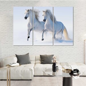 May include: Two white horses run through a snowy field. The horses are running side by side, with their manes flowing in the wind.