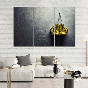 May include: A pair of gold boxing gloves hanging on a gray textured wall. The gloves are tied together with a black cord.