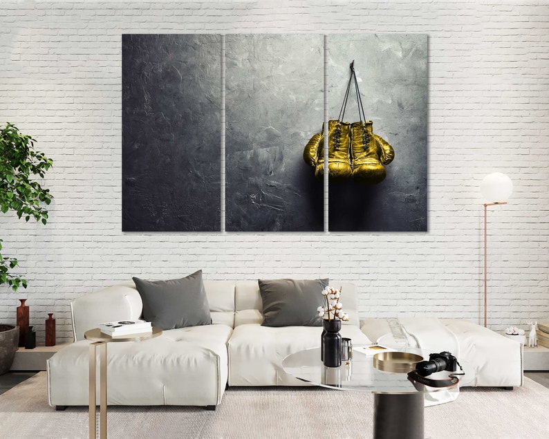 May include: A pair of gold boxing gloves hanging on a gray textured wall. The gloves are tied together with a black cord.