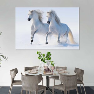 May include: Two white horses running through a snowy field. The horses are running towards the right side of the image. The snow is falling softly and the sky is a light blue.